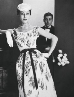 Bettina Graziani portant une robe fourreau brodée de motif floraux de Jacques Fath. Un homme derrière porte un bouquet de tulipes.